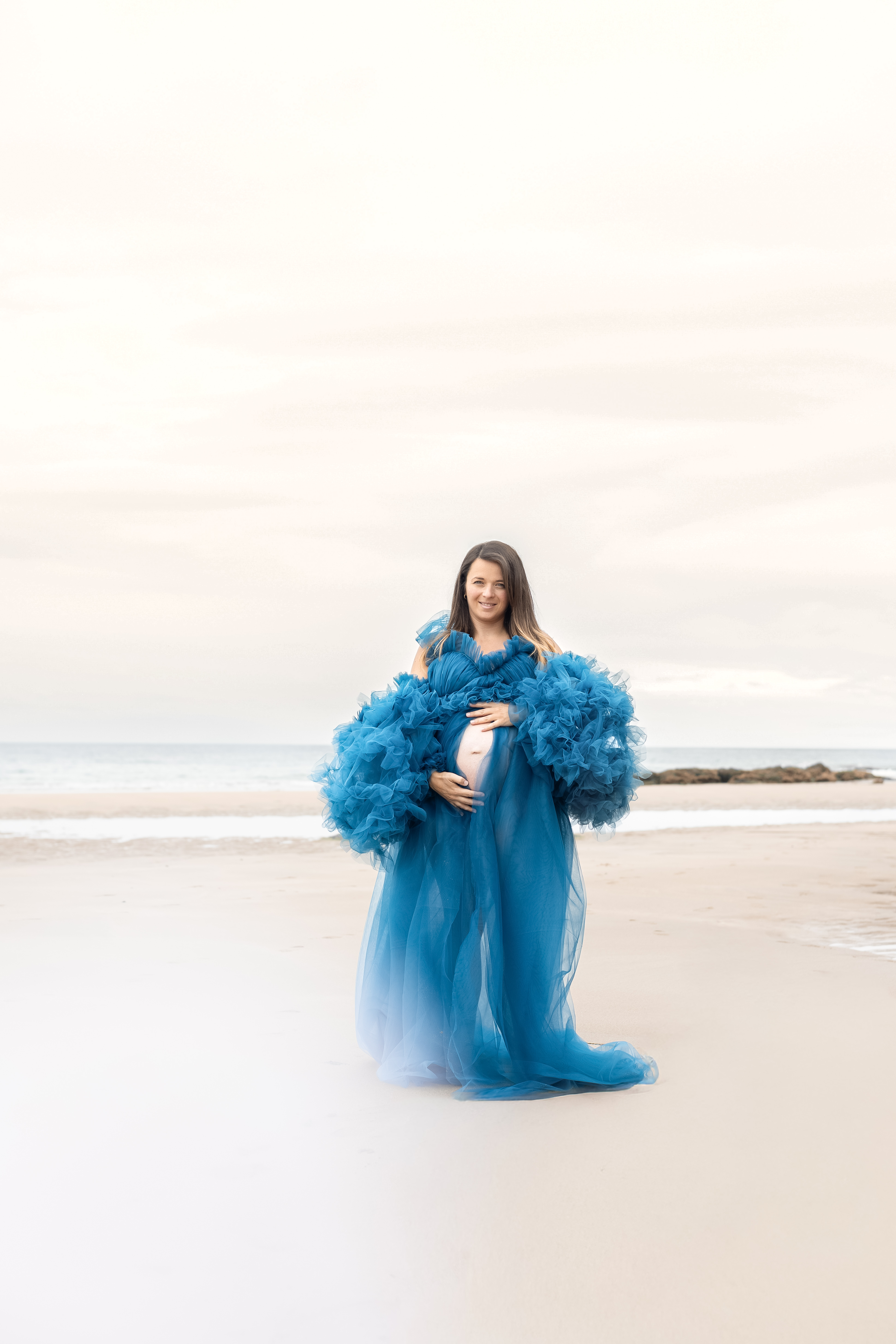 Future maman en robe bleue photographiée au loin sur une plage lumineuse